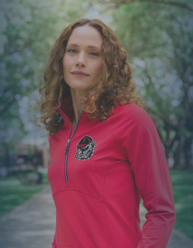 Woman wearing a bright red quarter-zip with a University of Georgia logo, standing on a tree-lined path in a sunny park.