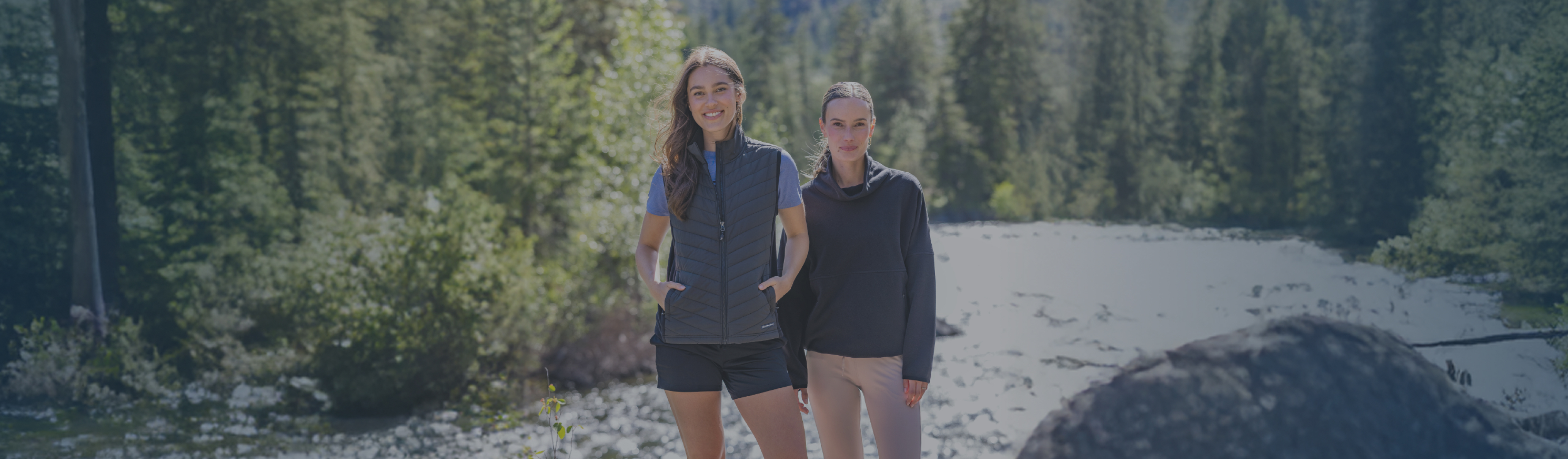 Two women standing on rocks beside a flowing river, wearing Cutter & Buck vests and pullovers. 
