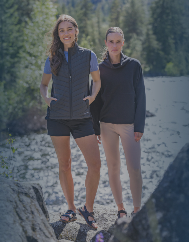 Two women standing on rocks beside a flowing river, wearing Cutter & Buck vests and pullovers. 