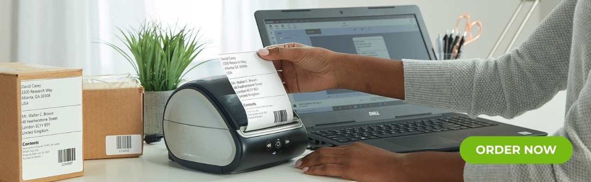 DymoOnline banner showing a desktop DYMO label printer producing shipping labels beside parcels, a laptop and office stationery to promote efficient, professional label printing.