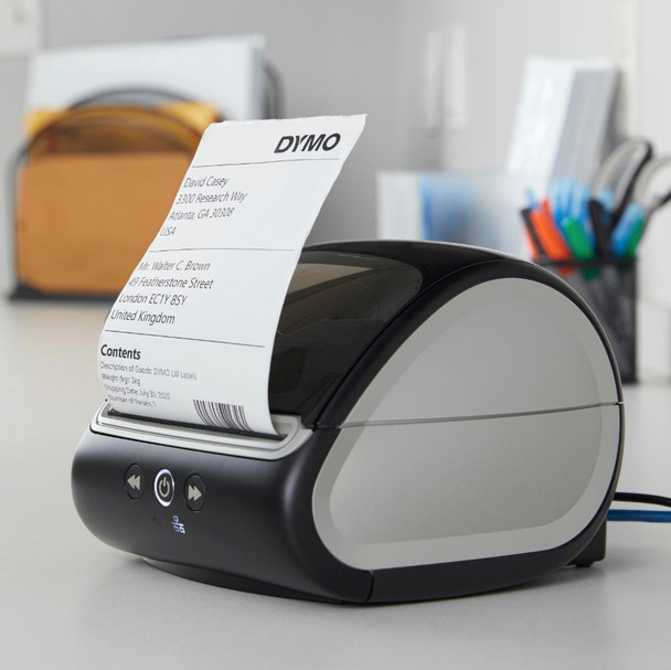 DYMO LabelWriter 5XL printer on a packing desk printing a shipping label beside boxes and a parcel scale.