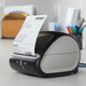 DYMO LabelWriter 5XL printer on a packing desk printing a shipping label beside boxes and a parcel scale.