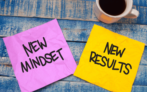 Sticky notes reading New Mindset and New Results on a blue wooden desk beside a coffee cup