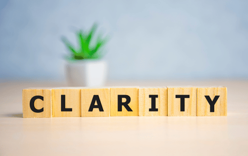 Wooden letter blocks spelling CLARITY on a desk with a blurred plant in the background