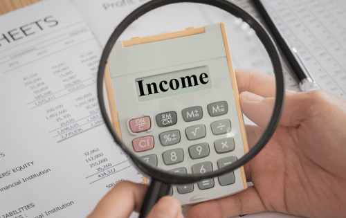 Hand holding a calculator under a magnifying glass with the word &ldquo;Income&rdquo; on the display, over financial documents.