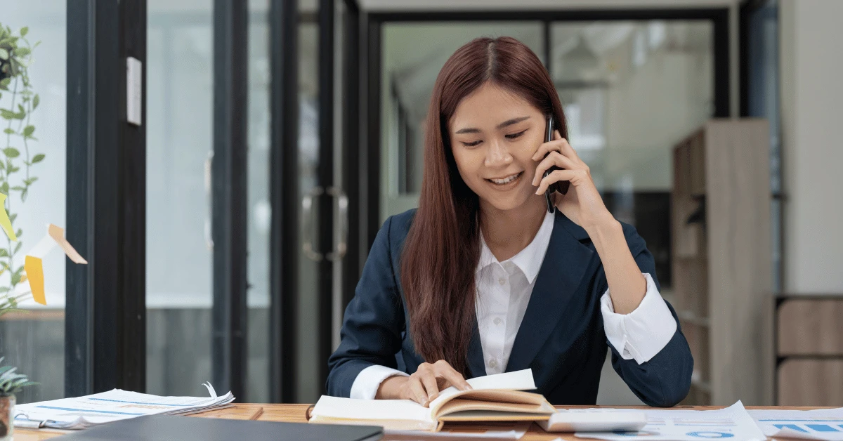 Businesswoman speaking on the phone while reviewing a notebook and documents at her desk.