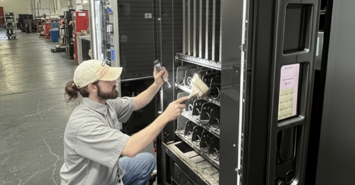Service technician cleaning and maintaining the inside of a vending machine in a warehouse, representing routine maintenance and equipment care for reliable vending operations