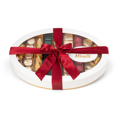 An elegant oval-shaped chocolate gift box with a matte gold base, crystal-clear lid, and festive red and green-wrapped chocolates, tied with a bright red bow and featuring the Mirelli logo.