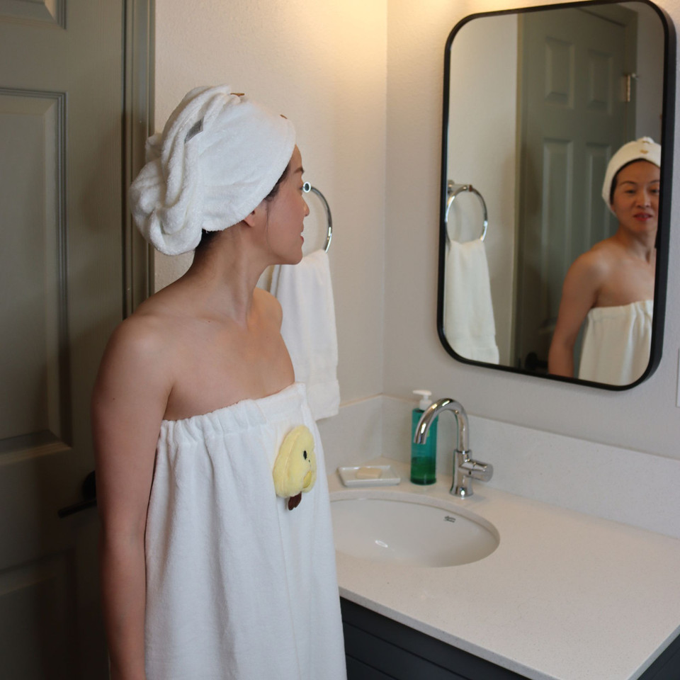 Woman wearing a white towel wrap set with a yellow character applique in a bathroom mirror