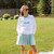 Woman stands on grass wearing a white sweatshirt that has the word birdie embroidered on the front of it in green