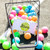 Basket of rainbow tennis balls tipped over on tennis court with white everyday pouch with lime green zipper is inside basket. Pouch has lime tennis ball with the word love in cursive printed on the front.