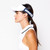 Woman stands on white background.  She is wearing a tennis ball overlap head in the game visor. Front of visor is white and sides are navy with white tennis balls printed on the sides.