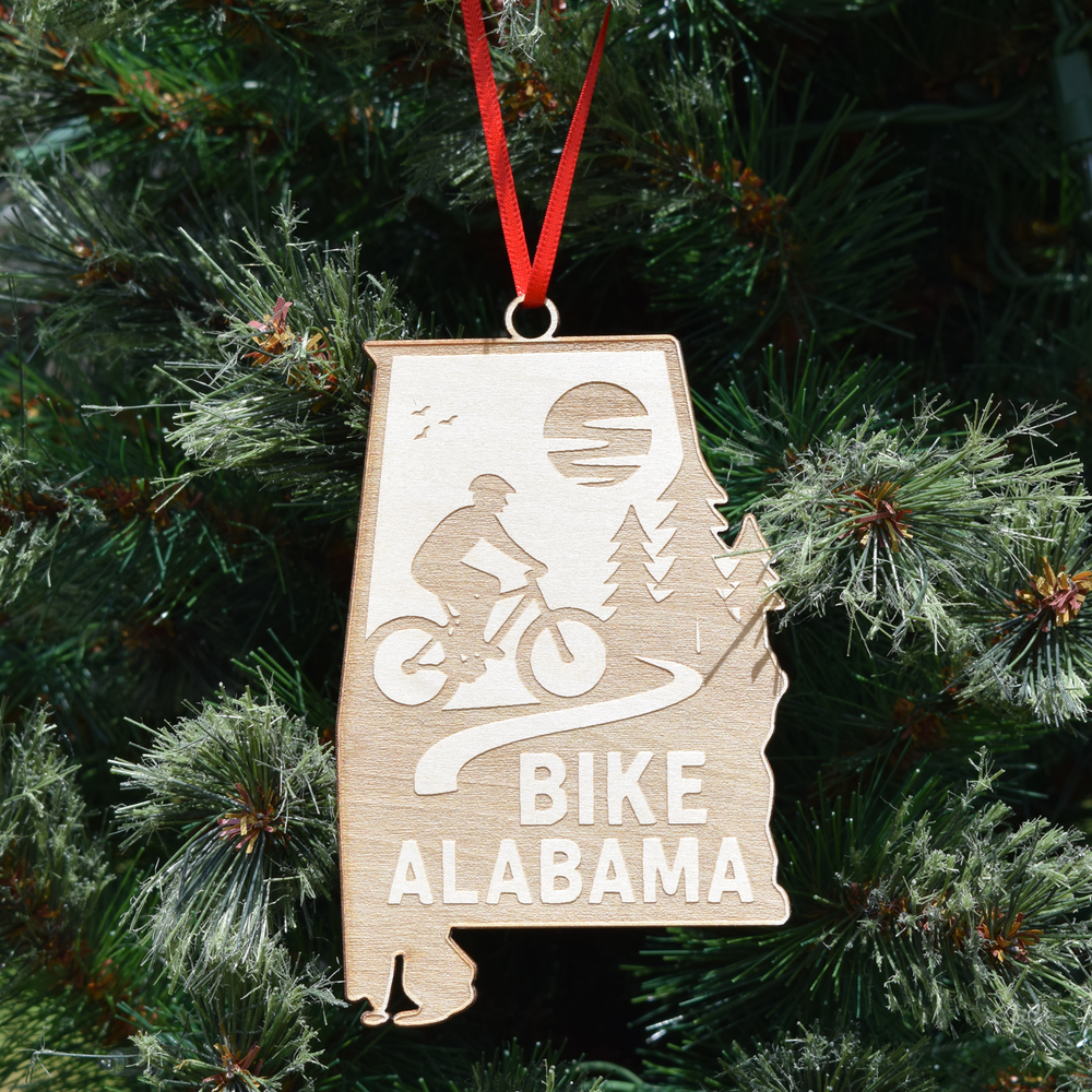 Bike Alabama wooden ornament shaped like the state of Alabama featuring a cyclist riding through a scenic trail landscape with laser-cut details and red hanging ribbon. (Ribbon Color May Vary)