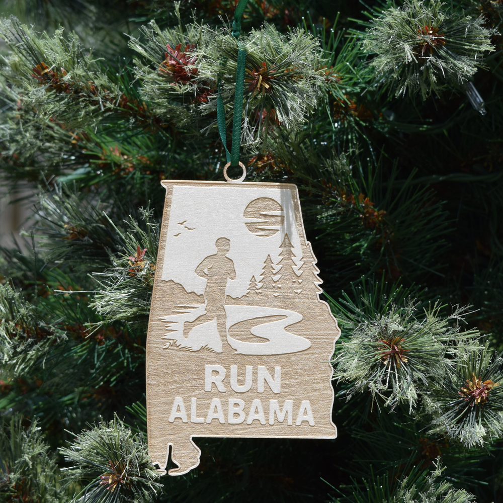 Run Alabama wooden ornament shaped like the state of Alabama featuring an engraved runner on a scenic trail with trees, natural wood finish, and hanging ribbon. (Ribbon Color May Vary)