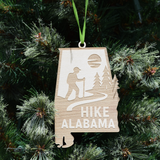 Hike Alabama wooden ornament shaped like the state of Alabama featuring an engraved hiker on a scenic trail with trees, natural wood finish, and hanging ribbon. (Ribbon Color May Vary)