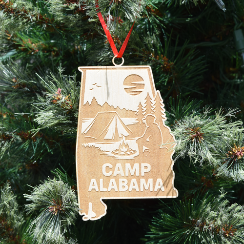 Camp Alabama wooden ornament shaped like Alabama featuring engraved camping scene with tent, campfire, trees, moon, and lake, hanging on Christmas tree.   (Ribbon Color May Vary)