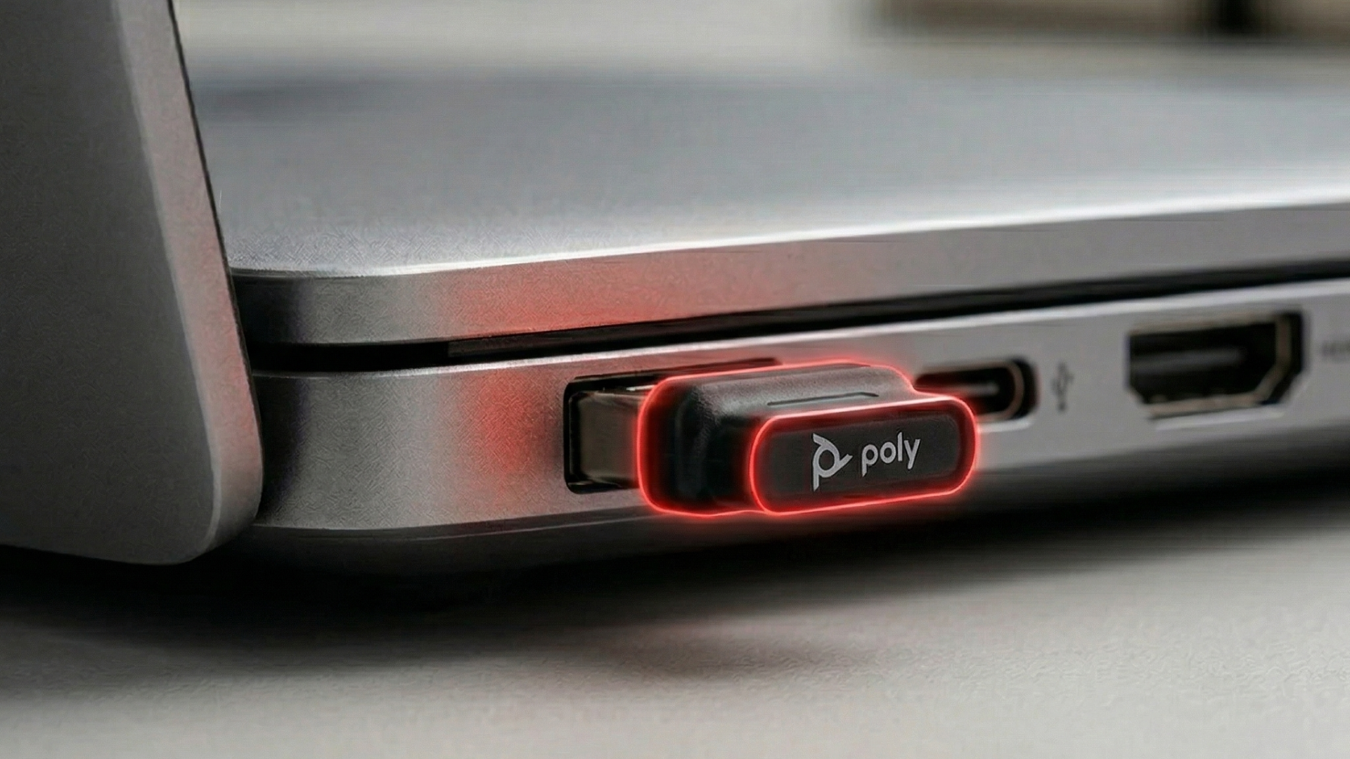 An extreme close-up of a dark gray Poly BT700 USB-A dongle plugged into the side port of a sleek, silver laptop. The dongle features the white 