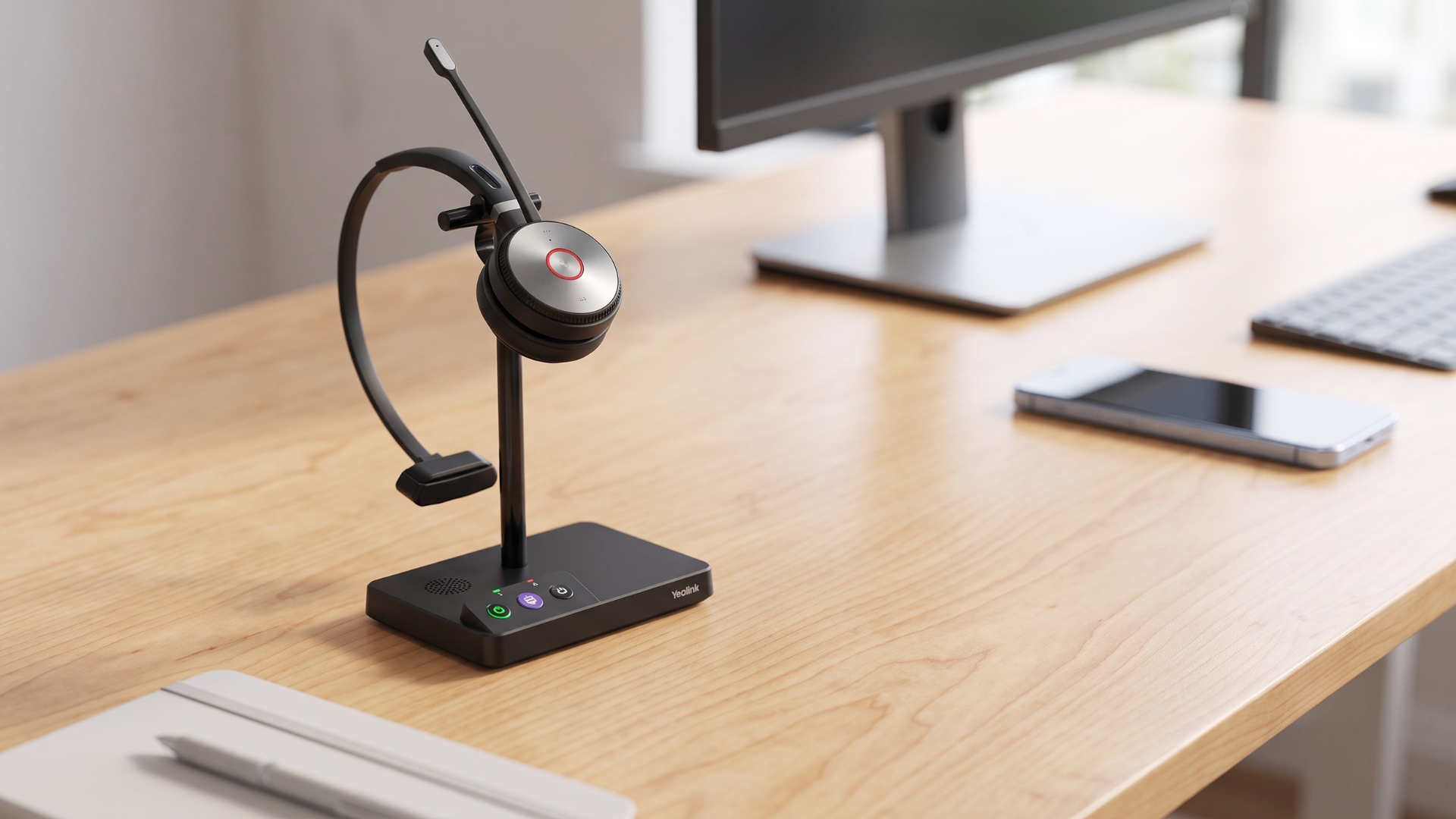 A clean photograph of a black Yealink WH62 mono wireless headset, with a silver-accented earcup and a boom microphone, docked on its charging base stand. The base sits on a light-wood office desk in a modern, naturally-lit environment. In the softly blurred background, a computer monitor, keyboard, and smartphone are visible. A notepad and pen are blurred in the foreground.