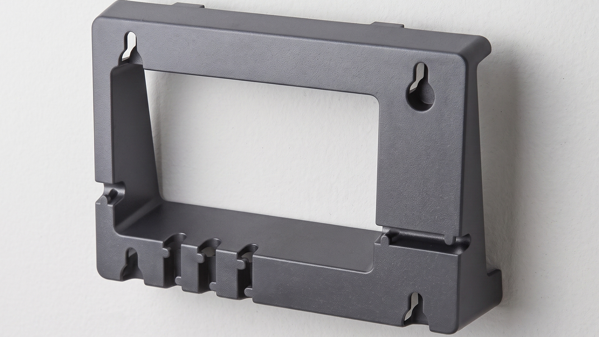 A detailed, photorealistic close-up view of the dark gray plastic Yealink WMB T46 wall mount bracket, as seen from a precise three-quarter quarter-view perspective. The bracket is securely installed on a light gray painted office drywall wall.