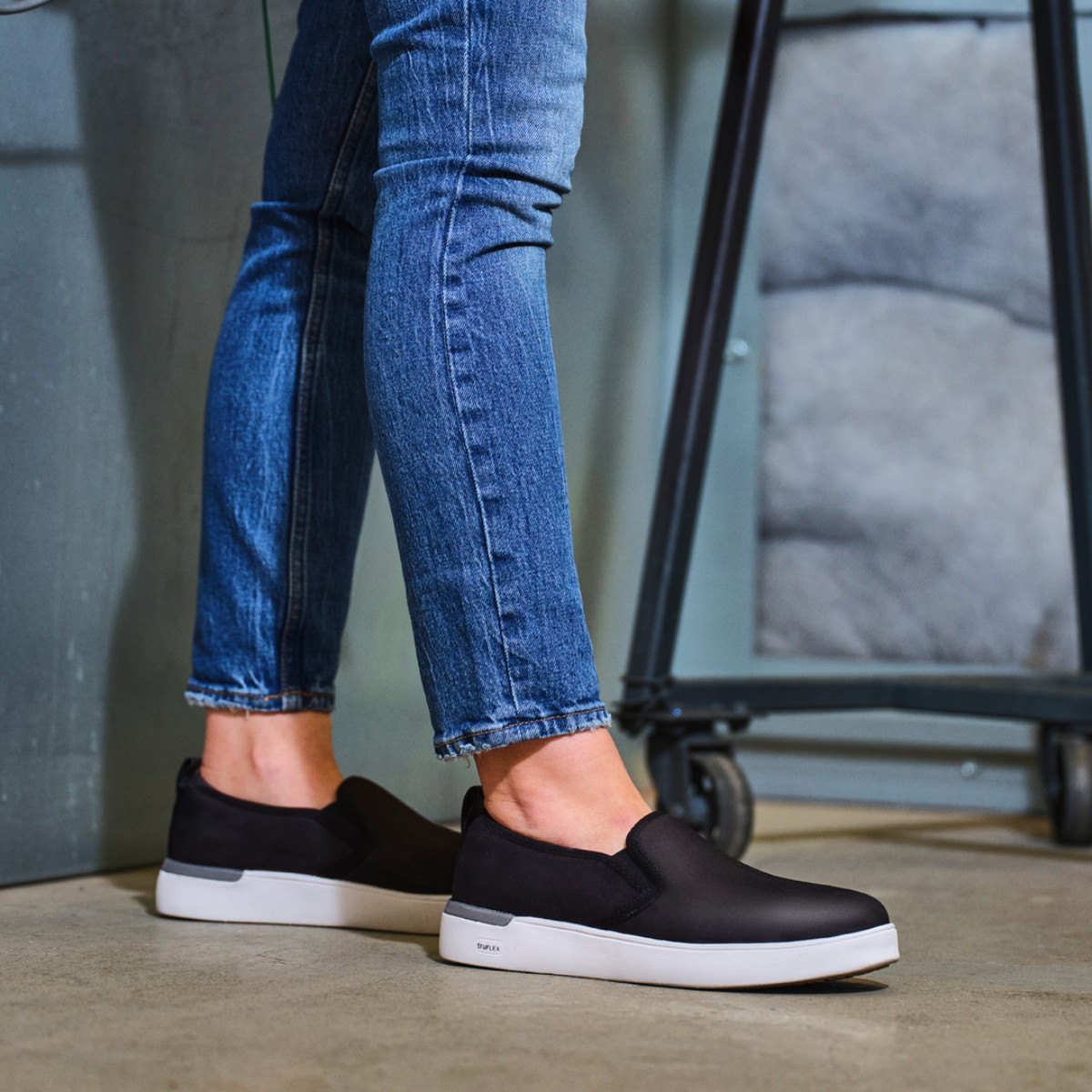 Parissa Work Women's Black Classic Work Slip-On - RK643 Person wearing black slip-on sneakers with white soles and blue jeans, standing on concrete floor