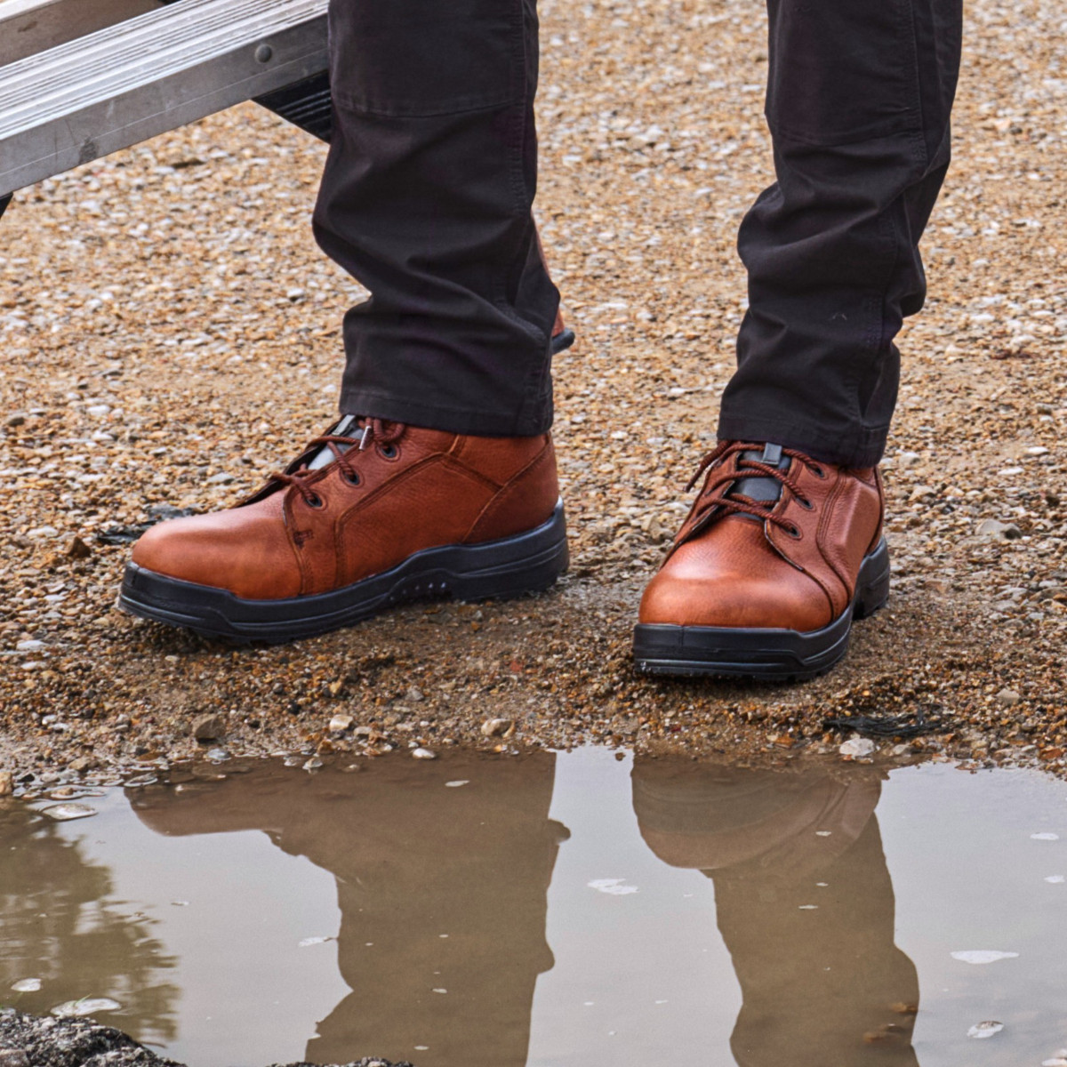 More Energy Men's Deer Tan 6 Inch Plain Toe Waterproof Work Boot - RK6628 Close-up of brown leather lace-up work boots with black soles on wet gravel ground, showing waterproof protection with puddle reflection