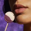Close-up of a woman holding a Sugar Splash DEEP Purple Razz Gag Suppressing Sucker near her lips against a purple galaxy background. The image highlights the sucker's distinctive purple-and-white candy design and reinforces its playful, oral-focused theme.