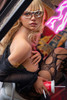 Close-up of Eileen IronTech silicone sex doll wearing glasses, reclining in black lingerie, showing her face, blonde hair and upper body.