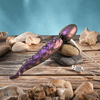 Iridescent purple tentacle vibrator resting across stacked stones, showing its flexible textured shaft, bulbous wand end and attached white USB charging cable.