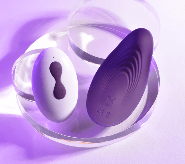 Playboy wearable panty vibrator with remote control displayed on a clear stand under purple lighting, highlighting sleek silicone design and discreet shape.
