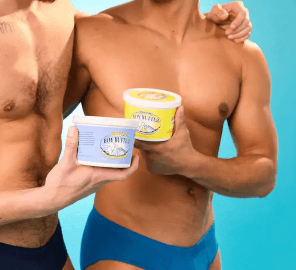 Two shirtless male models standing side-by-side against a light blue background, each holding a container of Boy Butter lubricant (one blue label, one yellow label). The focus is on the product in their hands with their torsos visible.