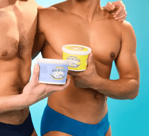 Two shirtless male models standing side-by-side against a light blue background, each holding a container of Boy Butter lubricant (one blue label, one yellow label). The focus is on the product in their hands with their torsos visible.