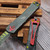 Detailed view of the Heretic Manticore S Bounty Hunter green distressed aluminum top scale with the deep-engraved red logo and MagnaCut blade.