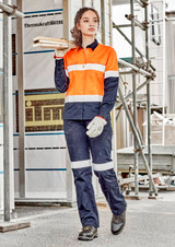 A woman in an orange and navy work shirt and matching pants stands on a construction site, holding wooden planks.