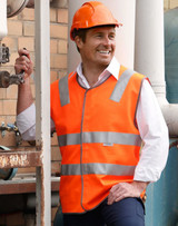 An orange hi-vis safety vest with reflective tapes, worn by a smiling man in a hard hat, stands near industrial equipment.
