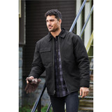 A black men's flatiron work jacket is worn by a man standing on stairs, holding gloves. The jacket features a pocket and has a casual style.
