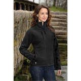 A woman stands by a stone pathway wearing a black soft shell jacket, featuring a zippered pocket and a logo.