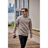 A man wearing a grey crew neck fleece sweater walks outdoors, holding a camera, with a city skyline in the background.