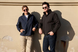 A navy ladies' soft shell jacket and a black men's soft shell jacket are worn by two models against a wall.