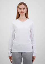 A woman wears a white long sleeve tee, paired with grey trousers. The tee features a simple design and has a logo.