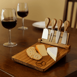 A wooden cheeseboard set featuring four stainless steel knives, cheese, and crackers, with two glasses of red wine in the background.