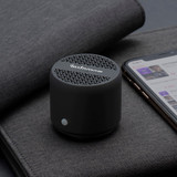 A compact black Bluetooth speaker rests on a grey surface next to a smartphone displaying a music app.