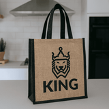 A jute tote bag in natural and black, featuring a lion design and the word "KING," with sturdy handles.