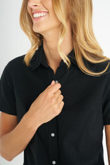 A black short sleeve shirt with a button-up front features a woman smiling while adjusting the collar.