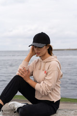 A woman wearing a black cap with a checker plate peak, paired with a beige hoodie and black pants by a waterfront.