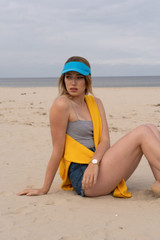 A yellow and blue visor sits on a model at the beach, wearing shorts and a grey top, with ocean in the background.