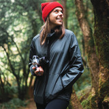 A woman wearing a dark green hooded rain jacket and a red beanie, holding a camera in a forest setting.