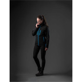 A woman wearing a black jacket with blue accents, styled with black leggings and boots, stands against a dark background.