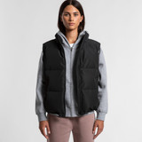 A black puffer vest worn over a grey hoodie, featuring a zip front and a logo. Model stands against a plain background.