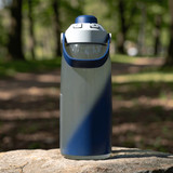 A drink bottle in grey and navy blue, featuring a large handle and a spout, placed on a rock in a natural setting.