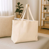 A cream-coloured reusable grocery tote bag with long handles, placed on a sofa. The setting includes natural decor elements.